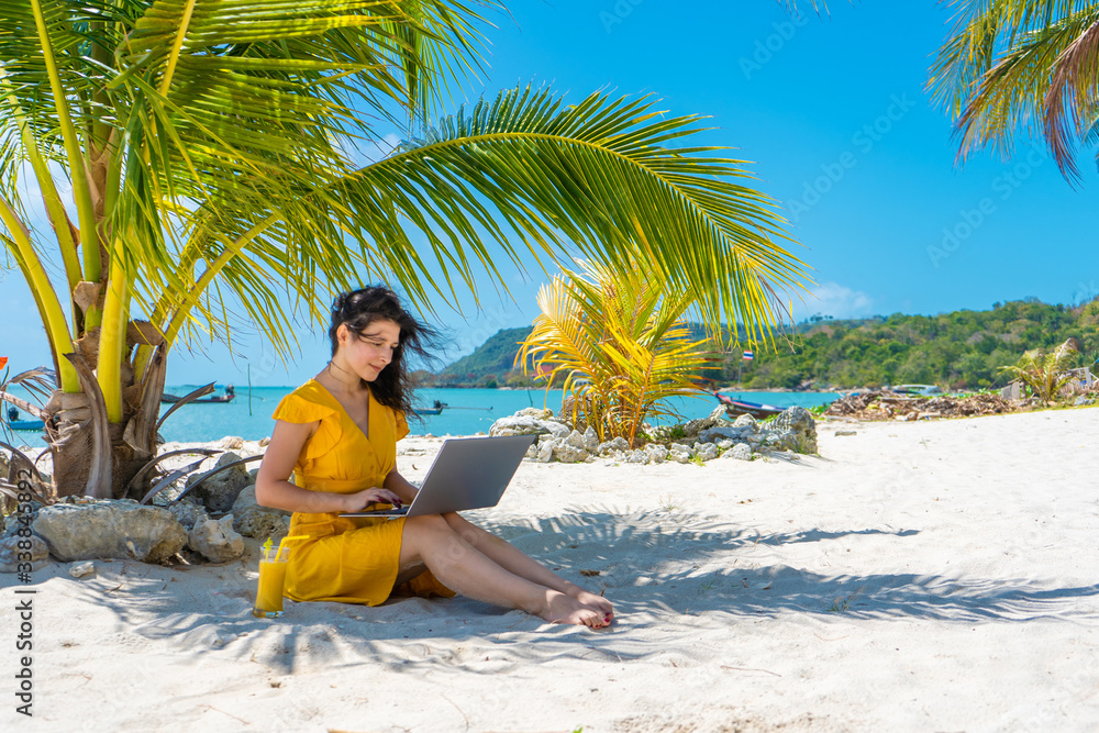 Girl in a yellow dress on a tropical sandy beach works on a laptop and drinks fresh mango. Remote work, successful freelance. Works on vacation.