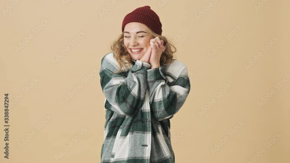 Pleased pretty woman in knit hat and shirt holding hands together and looking at the camera over beige background Pleased pretty woman in knit hat and shirt holding hands together and looking at the camera over beige background
