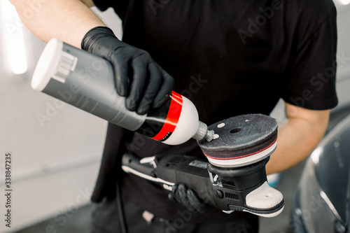Cropped image of auto service worker, wearing black t-shirt and gloves, putting special polish wax or cream on the orbital polisher to polish the car behind photo