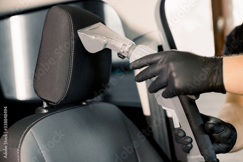 Cropped image of hands of car service worker, wearing black protective gloves, cleaning car front seat with wet vacuum cleaner. Car wet chemical vacuum cleaning, car detailing concept