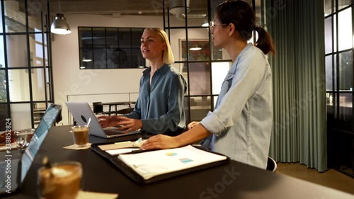 Two diverse young businesswomen talking to colleagues and smiling during a meeting around a table in an office