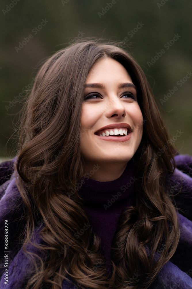 Girl smiling at camera on winter woods background. Glamorous funny young woman with smile wearing stylish sweater, purple fashionable fur coat. Fur and fashion concept. Beautiful people.