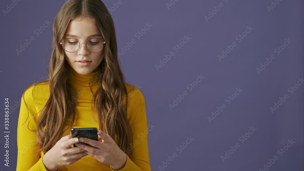 A calm nice teenage girl is using her smartphone standing isolated over a purple background in studio A calm nice teenage girl is using her smartphone standing isolated over a purple background in studio