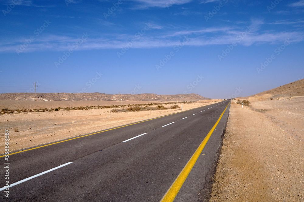 The desert landscape with the far yellow sandstone hills, the big empty black asphalt road, the blue sky with white clouds.