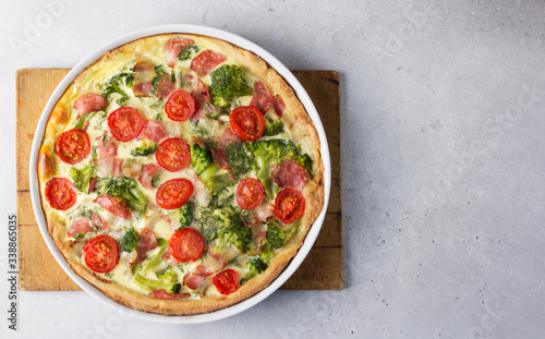 Open pie - quiche with bacon, broccoli, cherry tomatoes in white baking form on a wooden cutting board