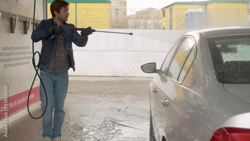 Handsome man washing car using high pressure washer. Self service on car wash. Russian text on the wall - administration is not responsible for damage resulting from improper use of equipment
