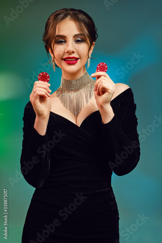 Brunette model in black dress, necklace and earrings. She is smiling, showing two red chips, posing on colorful background. Poker, casino. Close-up