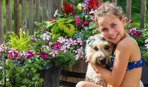 Young girl smiling while embracing a sunglassed wheaten terrier on her desk with many colorful flowers in the background. 