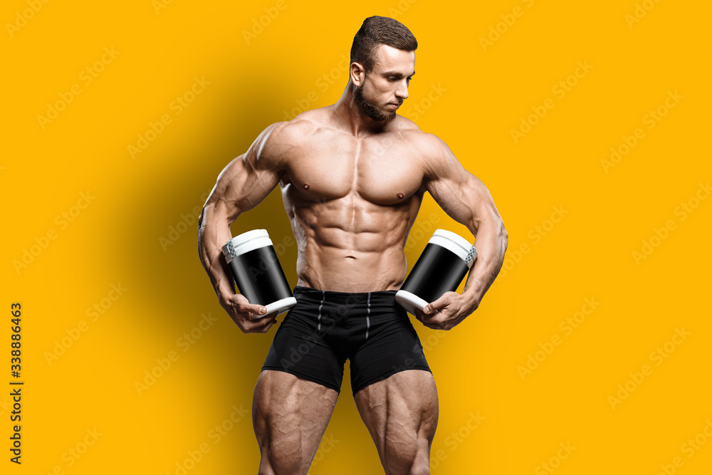 Muscular bodybuilder guy shows his perfect abs sixpack and holds sport nutrition supplement in brutal powerful hand isolated over yellow background. Workout bodybuilding concept.