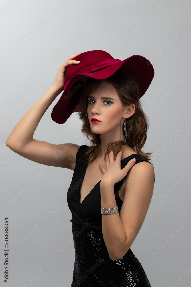Beautiful fashion photo of young girl with long curly hair in a black long dress and red hat posing on grey background. Concept of hairstyling in salon and face care Beautiful fashion photo of young girl with long curly hair in a black long dress and red hat posing on grey background. Concept of hairstyling in salon and face care