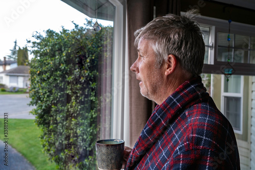 Unshaved middle aged man with messy unwashed hair holding coffee and looking out window in housecoat.