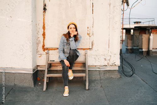 white European woman is sitting on a wooden staircase on the roof of a multi-storey building in stylish denim clothing and a Trendy yellow hat .in the rays of the setting sun. lifestyle concept.