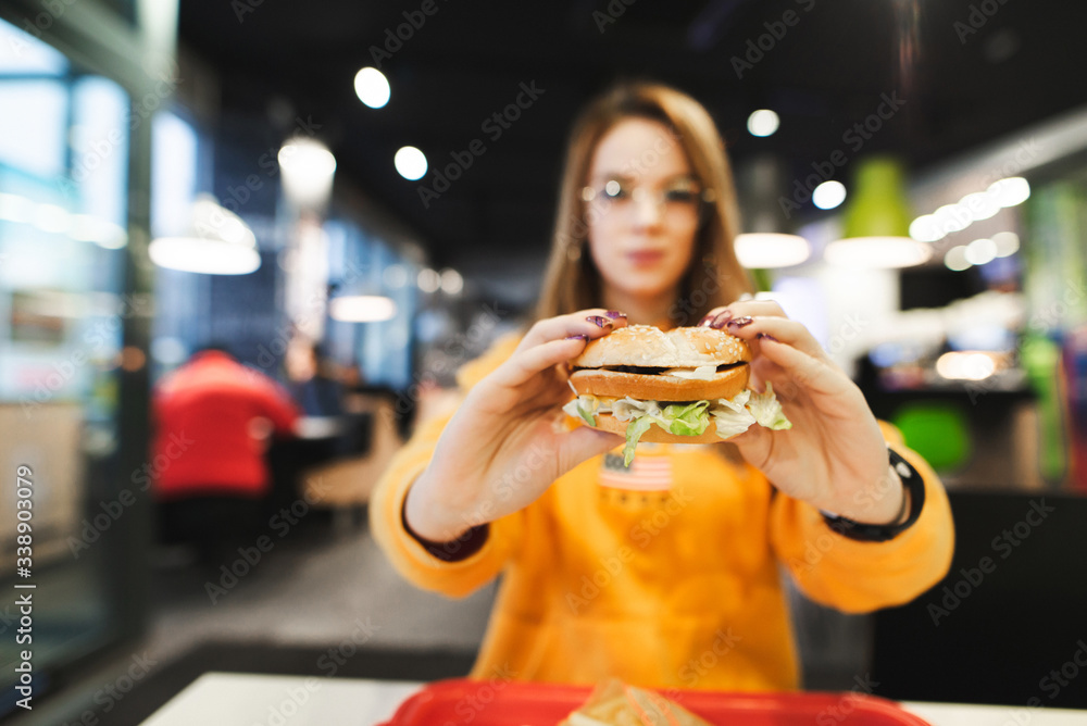 Girl sitting in cafe and holding burger in hand. Closeup photo of burger in hands of attractive girl having lunch in cafe. Background. Copy space
