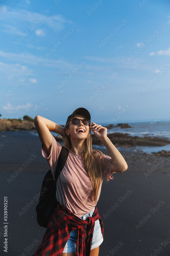 Vertical of happy attractive hipster woman traveller, backpack trip around Asia, enjoying Bali, Indonesia summer vacation, looking at sun standing on black volcanic sand contemplate blue ocean