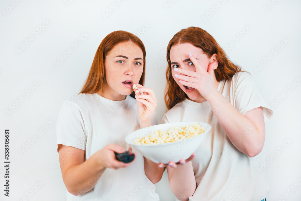 two redheaded scared women look frightened one with remote control another with popcorn standing on isolated white backgroung, leisure time concept two redheaded scared women look frightened one with remote control another with popcorn standing on isolated white backgroung, leisure time concept