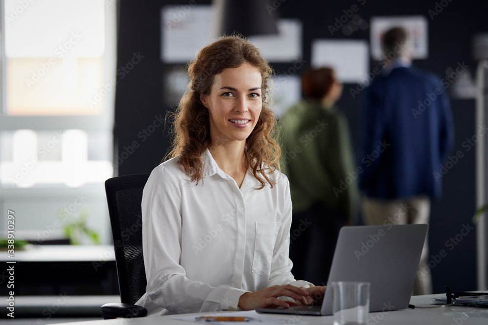Portrait of smiling businesswoman using laptop at desk in office with colleagues in background Portrait of smiling businesswoman using laptop at desk in office with colleagues in background