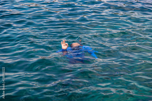 A man swimming underwater in the sea snorkeling  with a tube  in a blue suit  shows the OK sign