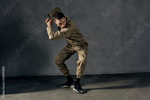 Graceful guy with tattooed body and face, earrings, beard. Dressed in khaki overalls, black sneakers. Dancing on gray background. Dancehall, hip-hop
