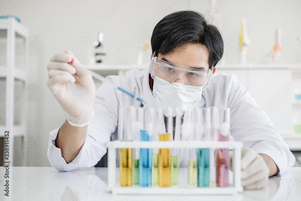 A male scientist with black hair wearing white coat and protective glassware and face mask dropping liquid solution from a pipet into a test tube looking serious in a laboratory setting. A male scientist with black hair wearing white coat and protective glassware and face mask dropping liquid solution from a pipet into a test tube looking serious in a laboratory setting.