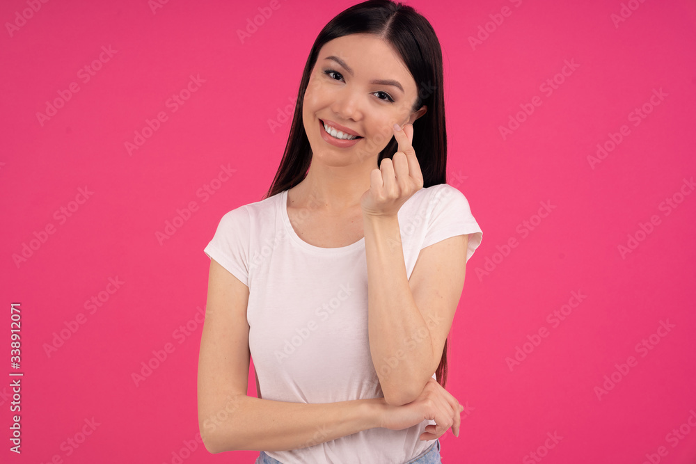Body language concept. Happy Asian woman makes korean hand sign, expresses love, makes like gesture, smiles gently at camera, dressed in casual wear, isolated on pink background. Monochrome. Body language concept. Happy Asian woman makes korean hand sign, expresses love, makes like gesture, smiles gently at camera, dressed in casual wear, isolated on pink background. Monochrome.