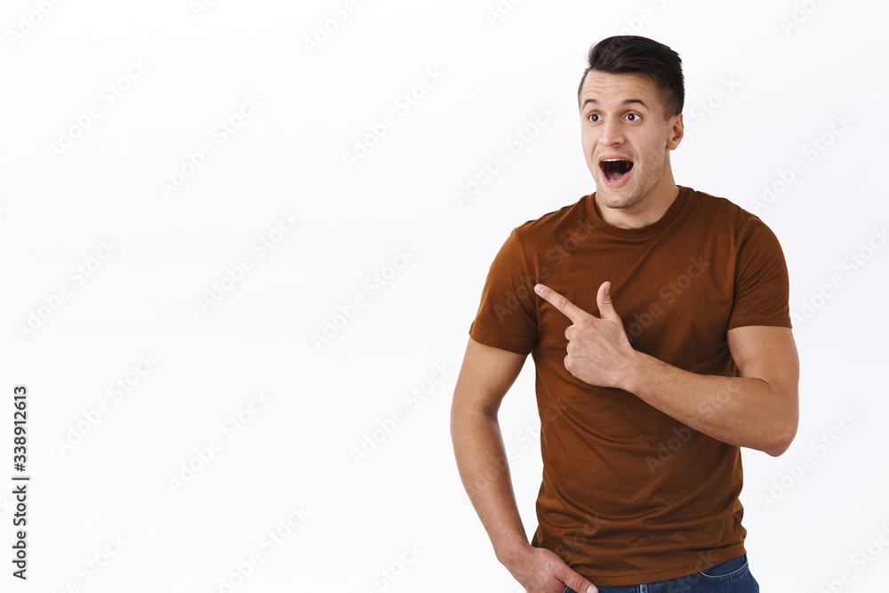Portrait of excited and happy caucasian male in brown t-shirt, open mouth fascinated looking and pointing fingers left, staring astonished as standing white background, seeing cool product Portrait of excited and happy caucasian male in brown t-shirt, open mouth fascinated looking and pointing fingers left, staring astonished as standing white background, seeing cool product