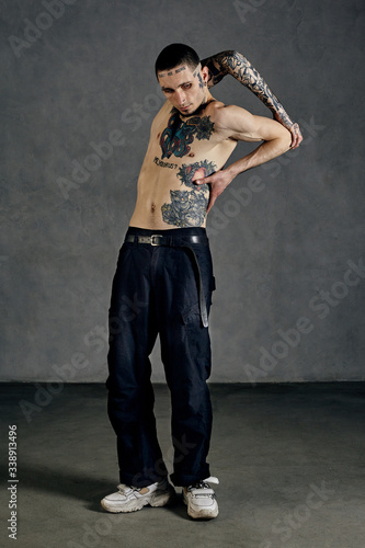 Flexible tattooed male with naked torso, beard. Dressed in black pants and white sneakers. Dancing against gray background. Dancehall, hip-hop