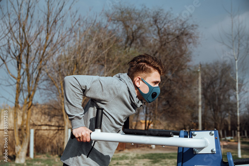 Young man in protective mask doing push-ups on uneven bars in outdoor street gym