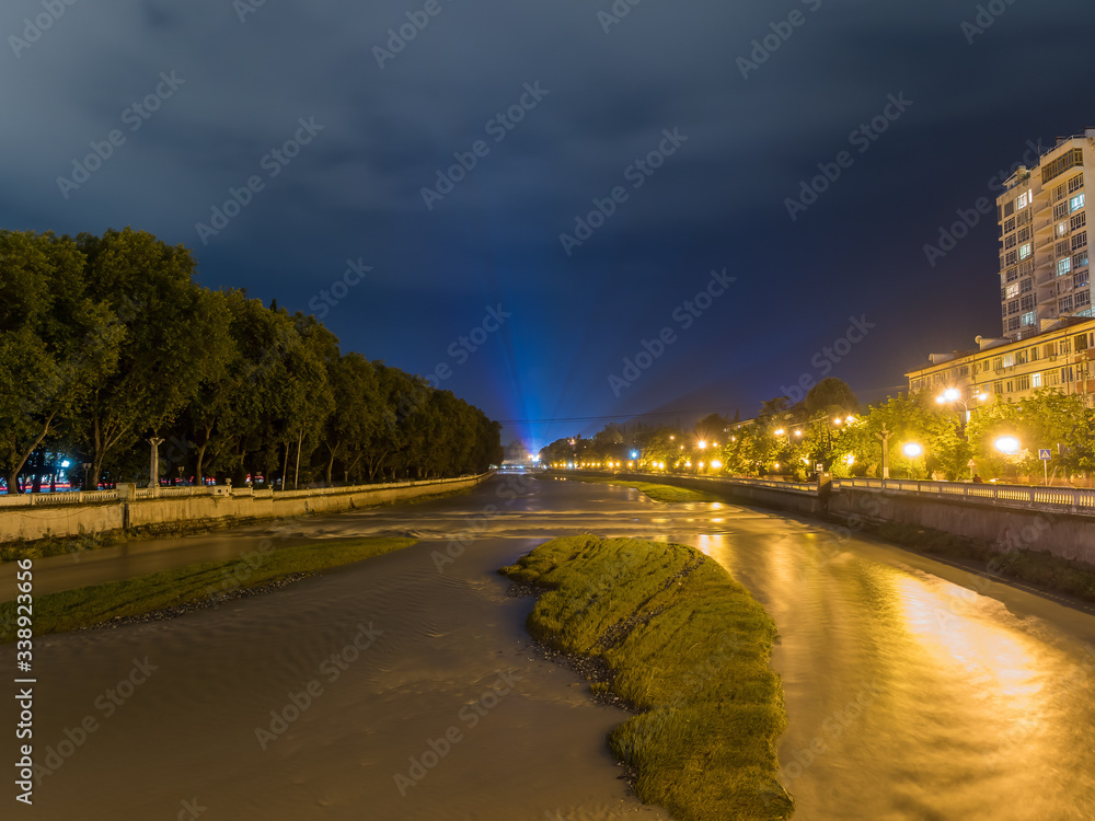 Night view of the wide city river illuminated by lanterns with rays in front with islands in the middle of the river and with trees and houses along