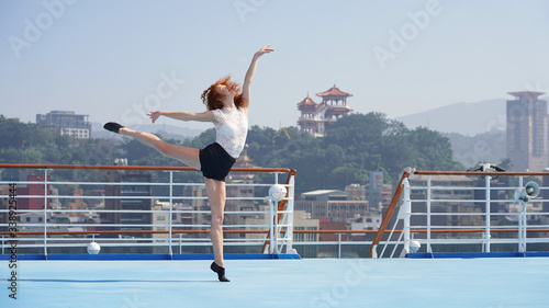 Elegant dancer. Beautiful slim sports model. Beautiful girl with long legs performs a dance pose balancing on one leg and toes. Woman on a cruise ship against the background of the Asian port city