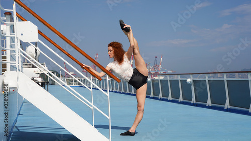 Flexible red-haired girl with beautiful legs standing on one leg performs a split. Graceful model on a blue background of a cruise liner in a cross split Elegant dancer. Beautiful slim sports model.