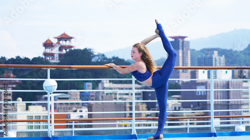 Beautiful athletic curly girl in bright clothes on deck of cruise ship performs gymnastic split. Flexible blonde acrobat smiles while standing in split against background of an Asian city, temple.