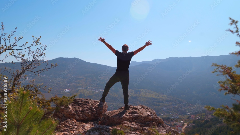 Young inspired man raises his hands up standing on the top of a mountain and looks at a beautiful valley of forest against beautiful blue sky. Silhouette of a happy man standing.
