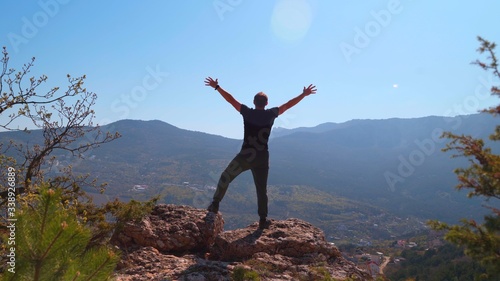 Young inspired man raises his hands up standing on the top of a mountain and looks at a beautiful valley of forest against beautiful blue sky. Silhouette of a happy man standing. © Nicolaos