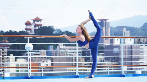 Beautiful slim European girl curly hair on observation deck of ship, against the background of Asian city. Blond gymnast standing on one leg in split on railing, sunny day.Professional circus acrobat