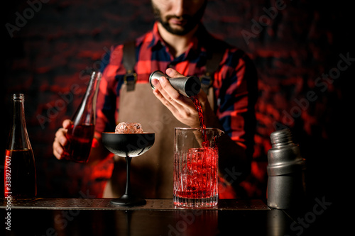 professional barman pours red liquid from jigger into mixing cup