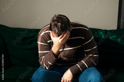 Frustrated young caucasian male burying face in hands sitting on green couch. Adult man showing signs of depression, anxiety, frustration and loneliness. Concept of breakdown and depression.