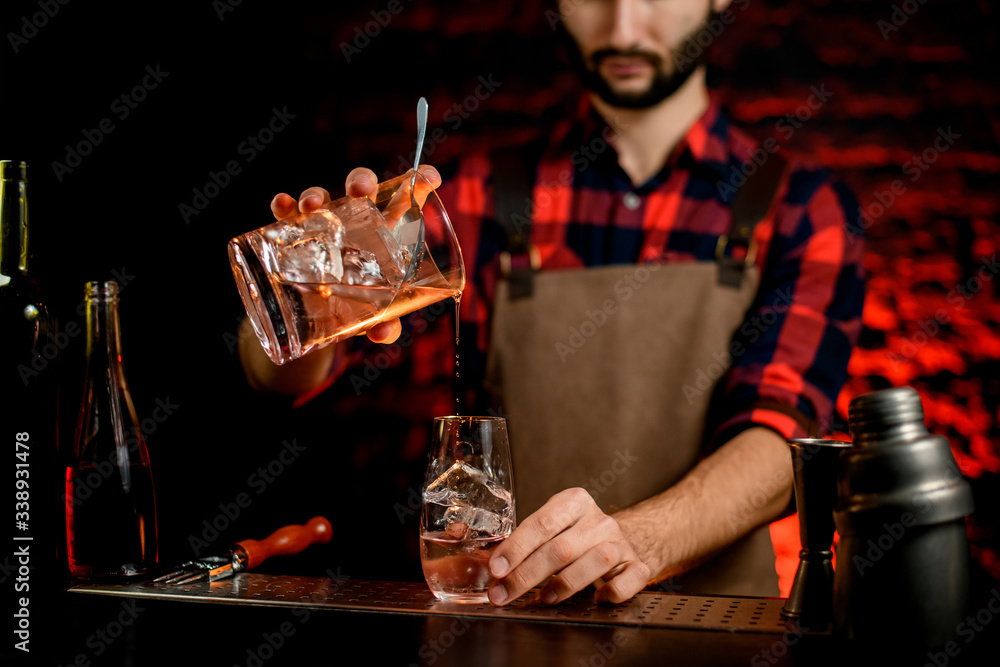 Man bartender professionally pours cocktail from mixing cup into small glass.