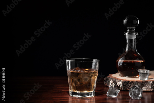 Crystal decanter or carafe of whiskey on cross section of birch tree trunk and glass with alcoholic drink and ice on wooden table in restaurant. Black background for copy space