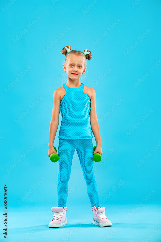Kid girl doing fitness or yoga exercises with dumbbells isolated on blue background