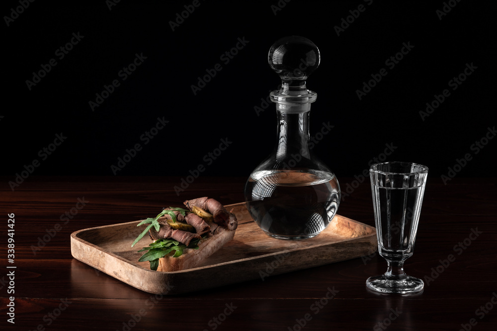 Crystal decanter or carafe with a glass full of vodka and tasty appetizer bruschetta with bacon and pickles on wooden board, alcohol drink concept with copy space Crystal decanter or carafe with a glass full of vodka and tasty appetizer bruschetta with bacon and pickles on wooden board, alcohol drink concept with copy space