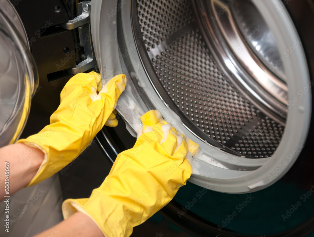 Female hands in yellow latex gloves for cleaning with a sponge wash the dirt around the drum of a black washing machine Female hands in yellow latex gloves for cleaning with a sponge wash the dirt around the drum of a black washing machine