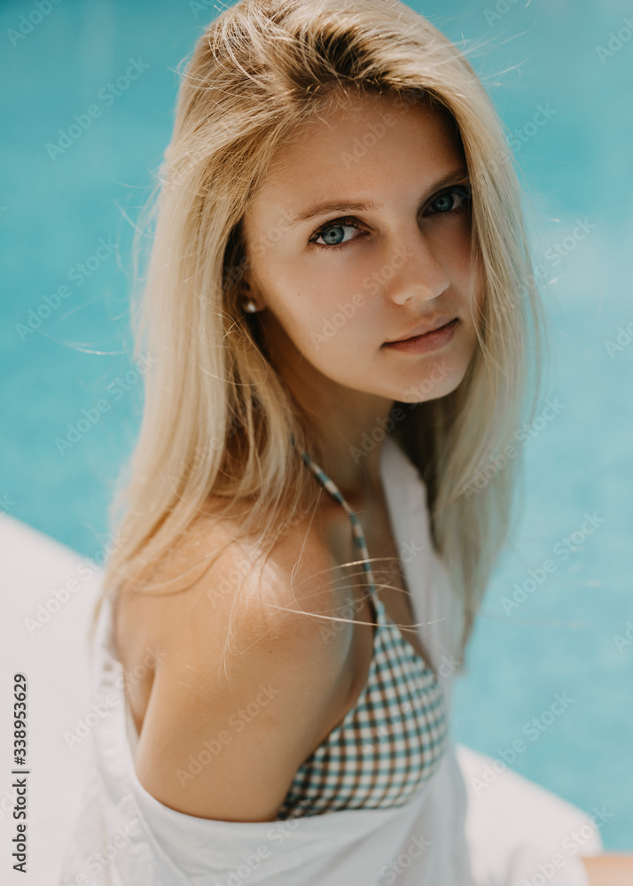 Close-up portrait of a young woman with blonde hair, wearing white shirt and bikini set, sitting by the pool. Close-up portrait of a young woman with blonde hair, wearing white shirt and bikini set, sitting by the pool.
