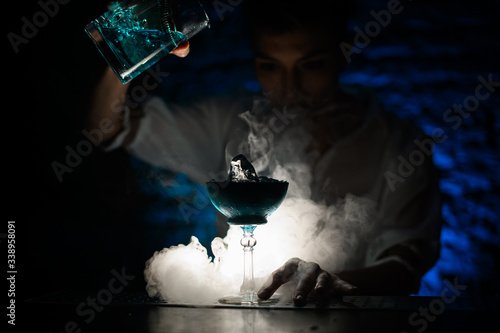bartender pours blue cocktail from mixing cup into glass at dark smoky bar.