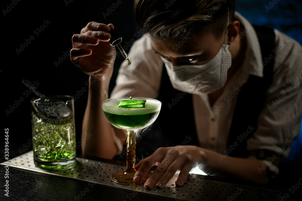 Woman bartender in mask carefully add ingredient to smoky glass with green cocktail.