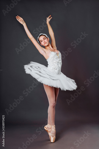 Portrait of young beautiful graceful caucasian ballerina practice ballet positions in tutu skirt of white swan from Swan Lake. Classical Ballet dancer in studio on gray background standing on tiptoe