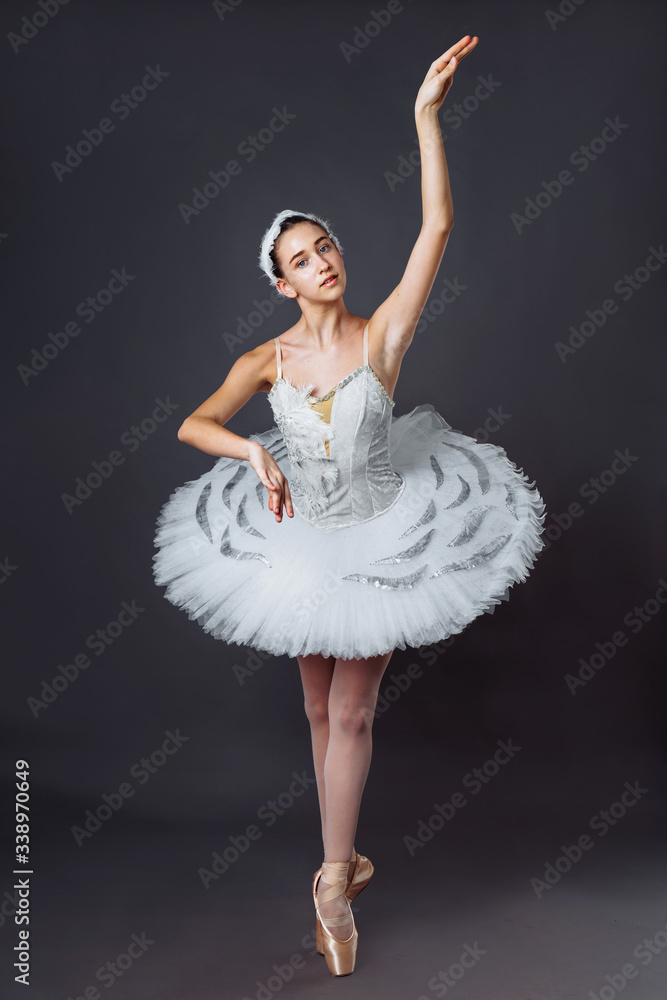 Fototapeta premium A young graceful ballerina dressed in professional attire, pointe shoes and a white tutu, demonstrates dance skills. Beautiful classic ballet dancer isolated on grey backgroung in photostudio
