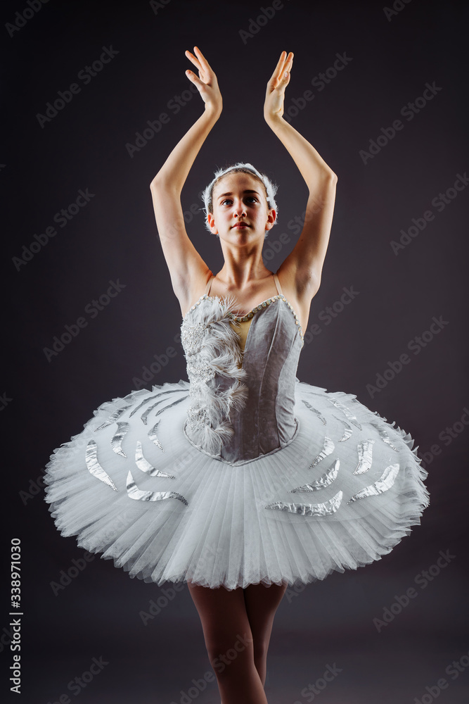 Fototapeta premium Portrait of the ballerina in the role of a white swan on grey background. Beautiful, attractive, young, graceful girl professionally performing ballet pas, dressed in a swan costume and pointe shoes