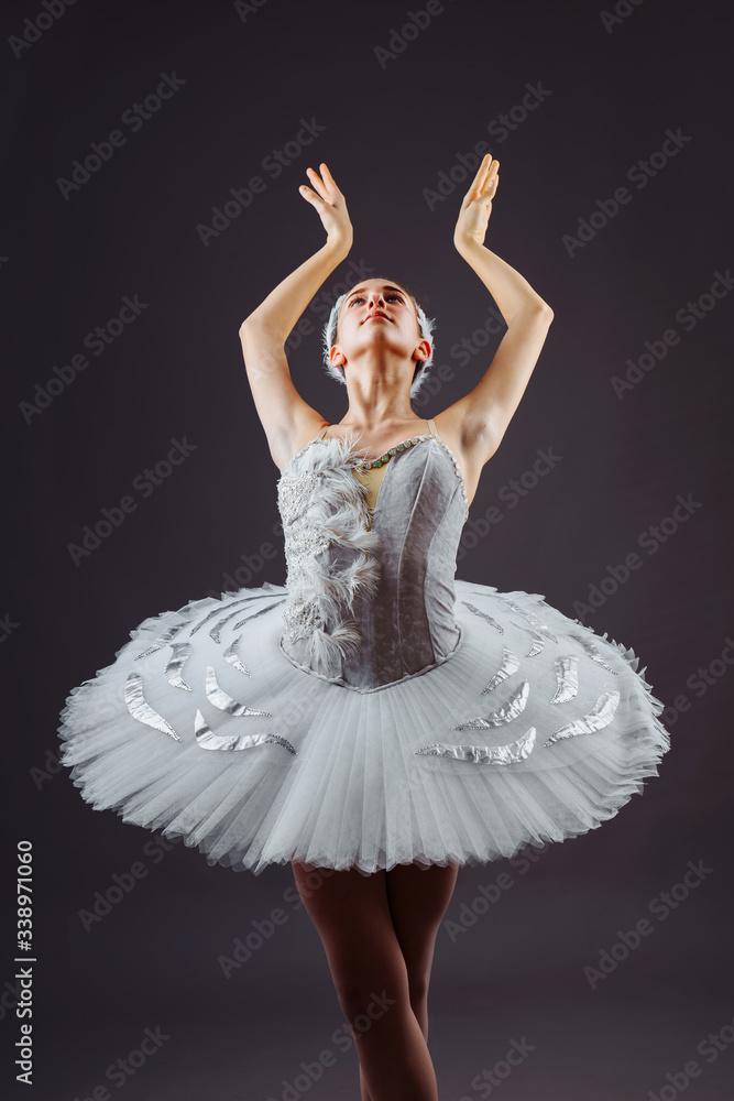 Fototapeta premium Portrait of the ballerina in the role of a white swan on grey background. Beautiful, attractive, young, graceful girl professionally performing ballet pas, dressed in a swan costume and pointe shoes
