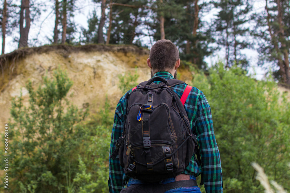 Handsome man hipster with beard on serious face in cloth shirt and suspenders sunny outdoor on mountain top against cloudy sky on natural. Tourism concept. Travel with a backpack. Wonderful landscape.