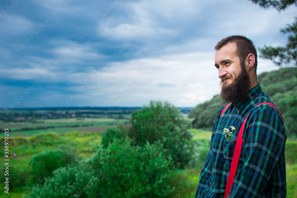Handsome man hipster with beard on serious face in cloth shirt and suspenders sunny outdoor on mountain top against cloudy sky on natural. Tourism concept. Wonderful landscape. Handsome man hipster with beard on serious face in cloth shirt and suspenders sunny outdoor on mountain top against cloudy sky on natural. Tourism concept. Wonderful landscape.